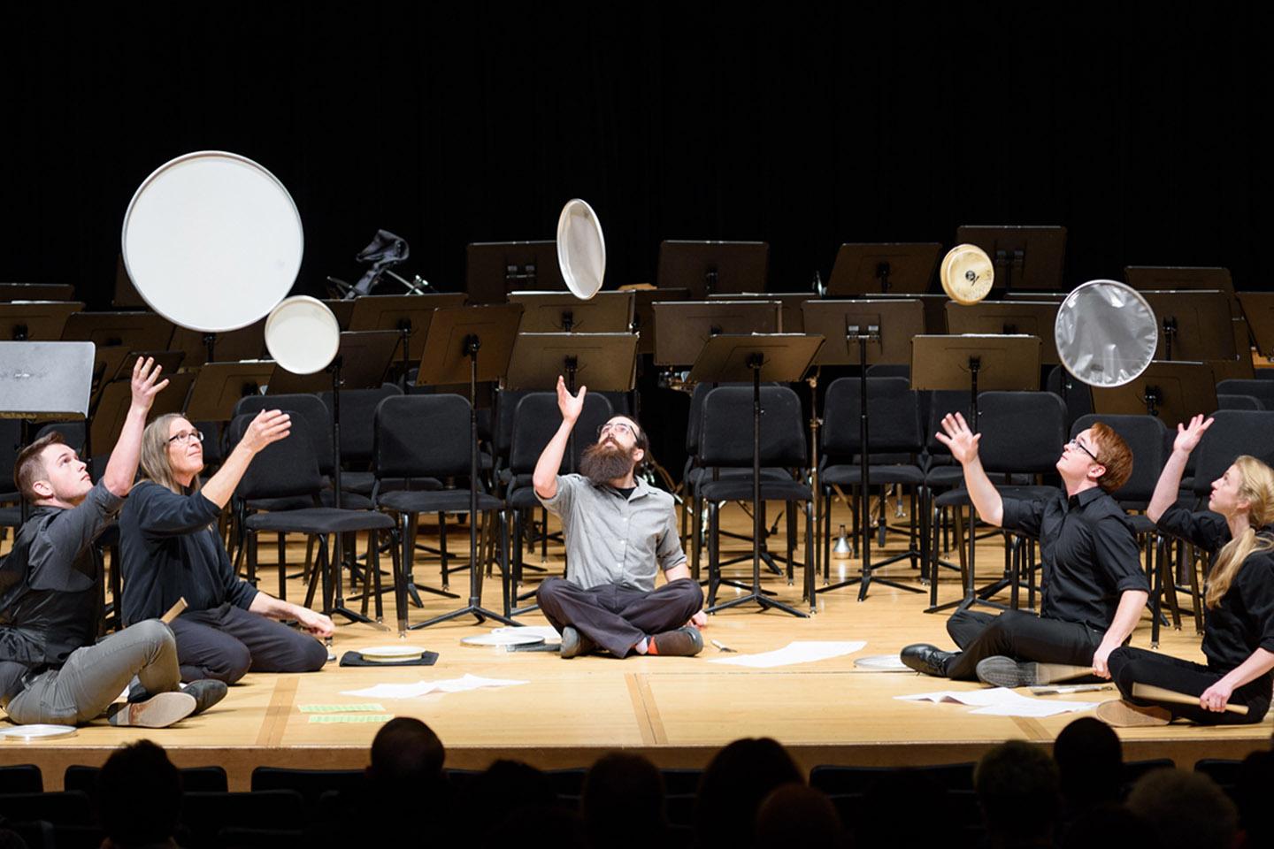 Students in percussion ensemble on stage throwing frame drums above their heads