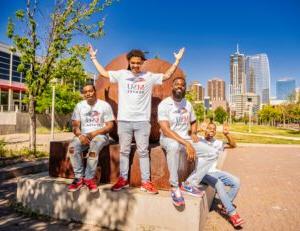 Photo of the first three MSU Denver MISTERs and the Call Me MISTER Director, Rashad Anderson, wearing matching MSU Denver t-shirts, posing on a sculpture outside on Auraria Campus with downtown Denver in the background.