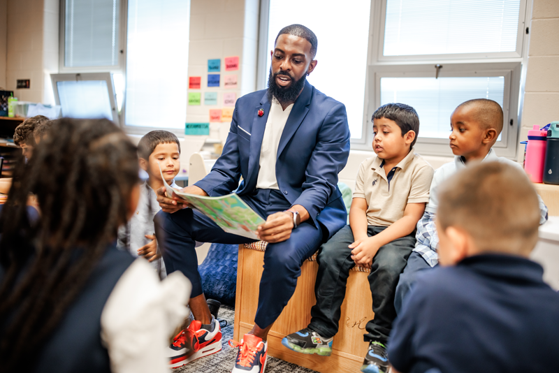 Dr. Rashad Anderson, Call Me MISTER program director, sitting on a bench in an elementary classroom reading to a small group of young students.