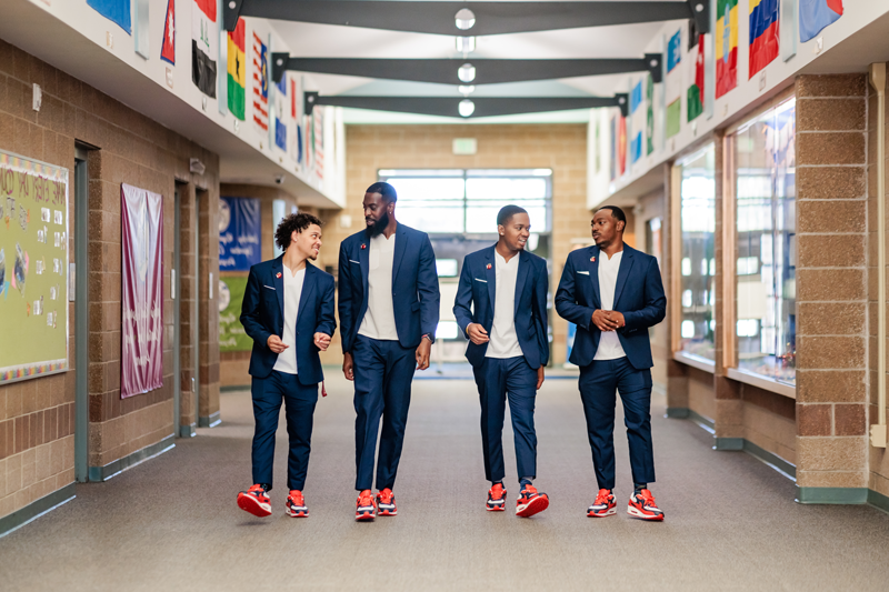 Call Me MISTER students Chris Livingston, Josh Barringer and Jordan Puch, along with Call Me MISTER program director Dr. Rashad Anderson, walking down the hallway of an elementary school toward the camera, wearing matching suits and sneakers, talking to one another.